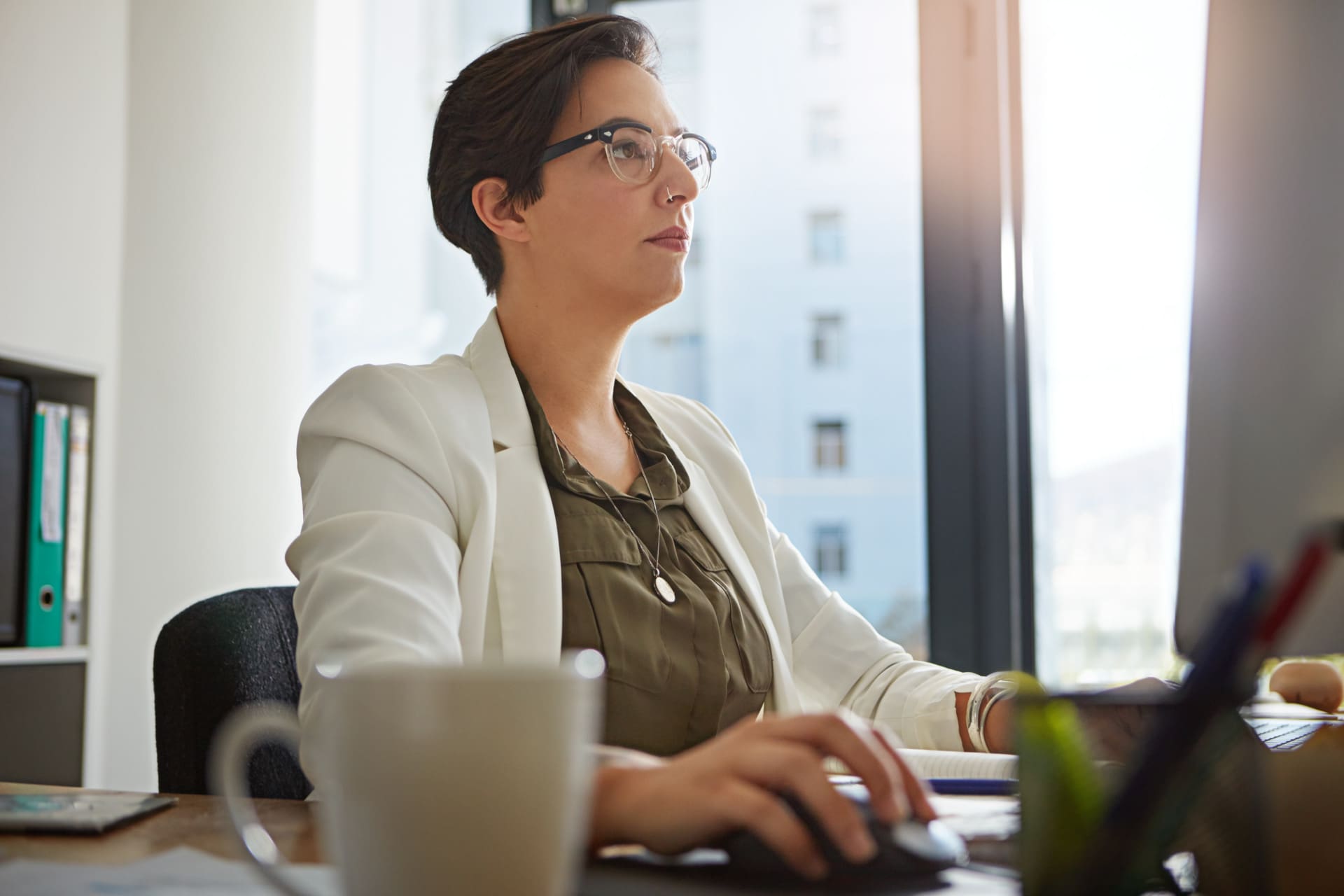 A person with short dark hair and glasses sits at a desk using a computer, possibly operating a Virtual Recorder. They wear a white blazer and olive shirt. A coffee mug sits in the foreground, with buildings visible through the window behind them.