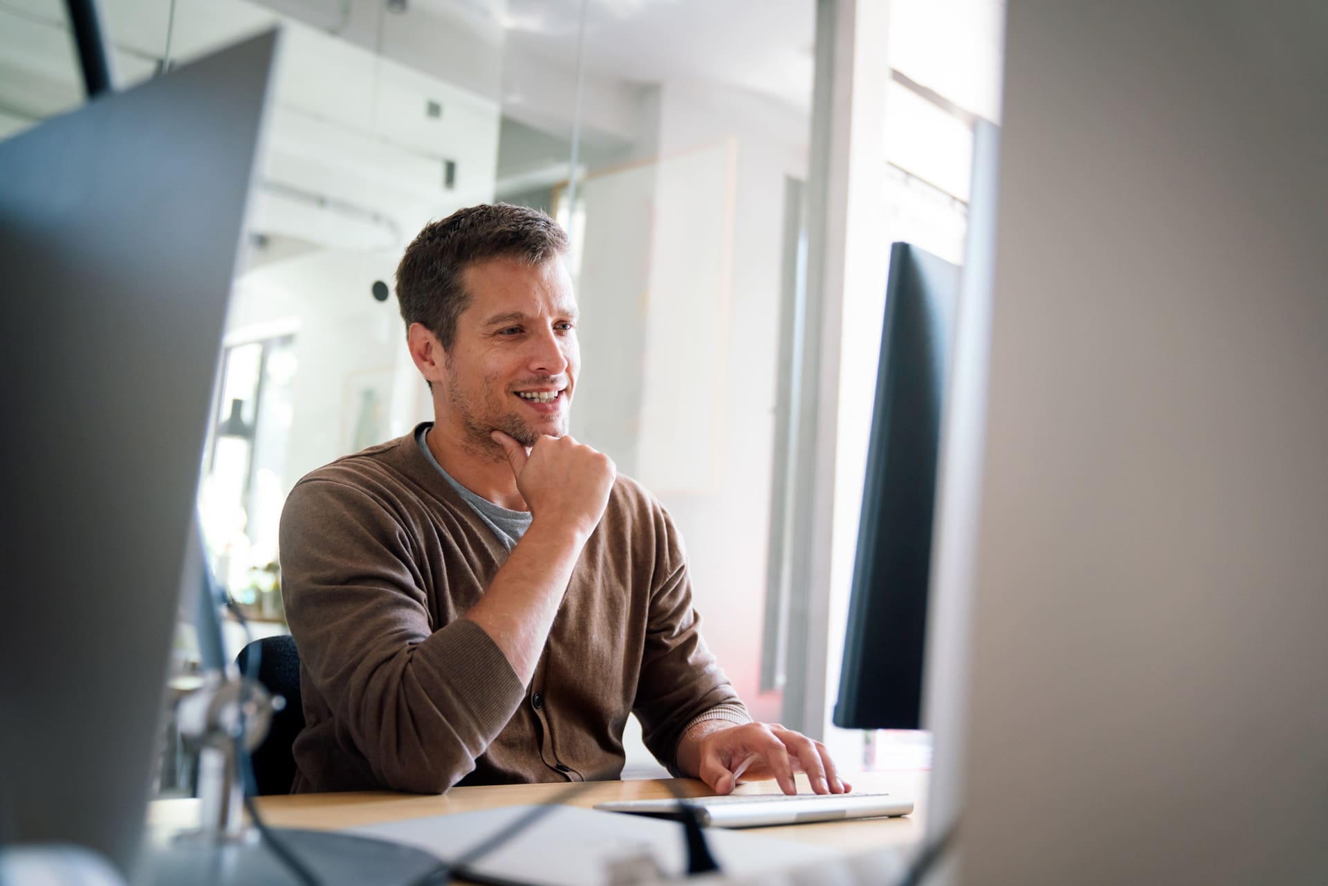 A man sits at a desk in an office, smiling while looking at a computer monitor. He rests his chin on one hand and uses the other hand to operate a computer mouse.