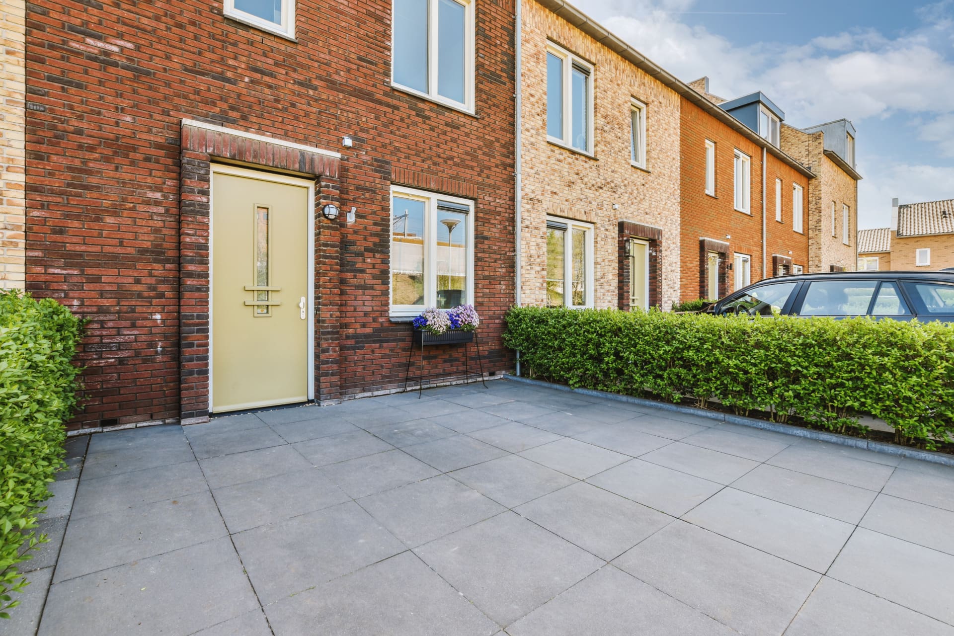 A row of modern brick townhouses with large windows and beige doors, featuring a small paved front yard, green hedges, and a parked car on the right side—ideal for those seeking their next home with PropertyWatch.
