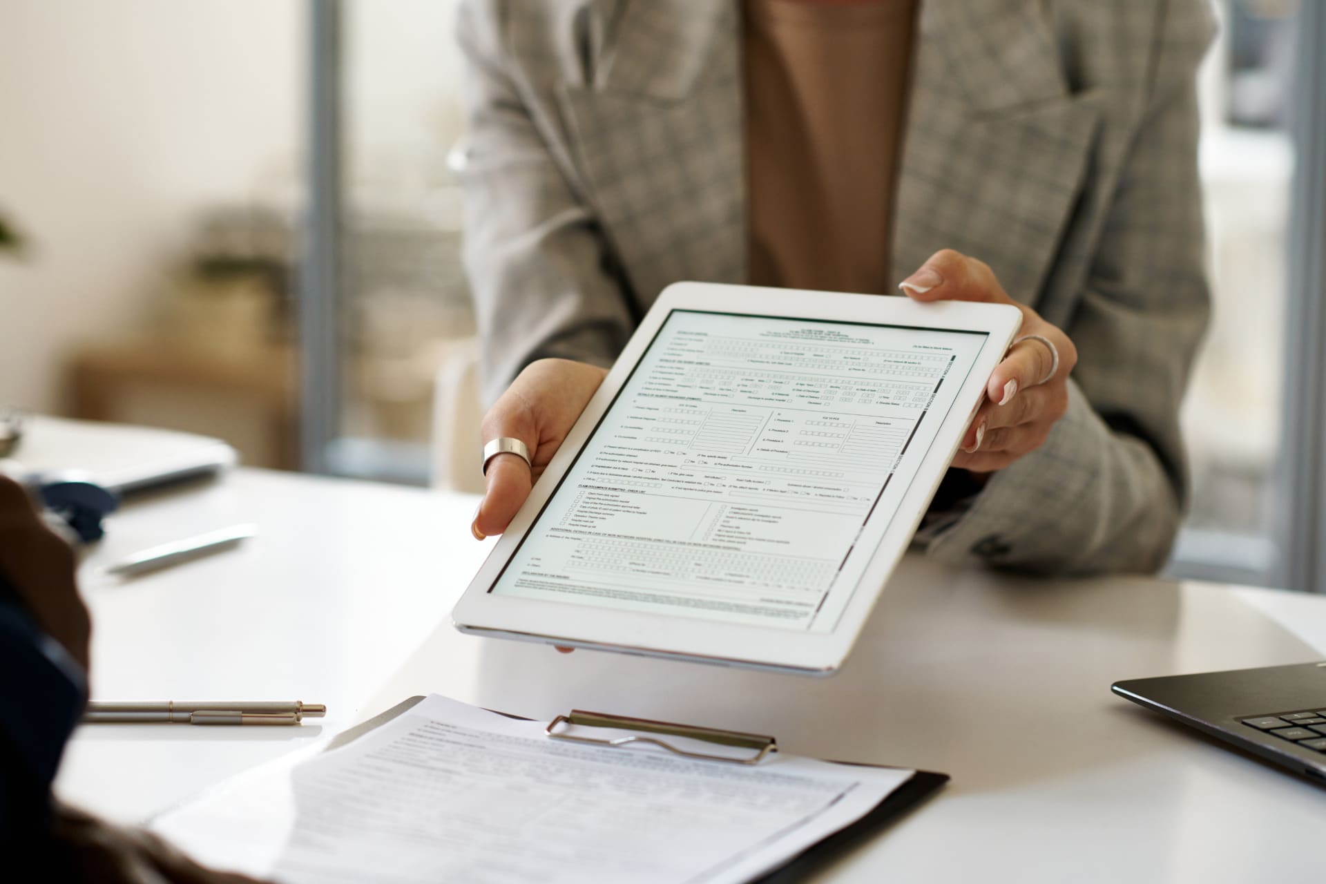 A person in a plaid blazer holds a tablet displaying an electronic document to another person across a desk, which has documents, a clipboard, a pen, and a laptop—highlighting seamless electronic document receiving.