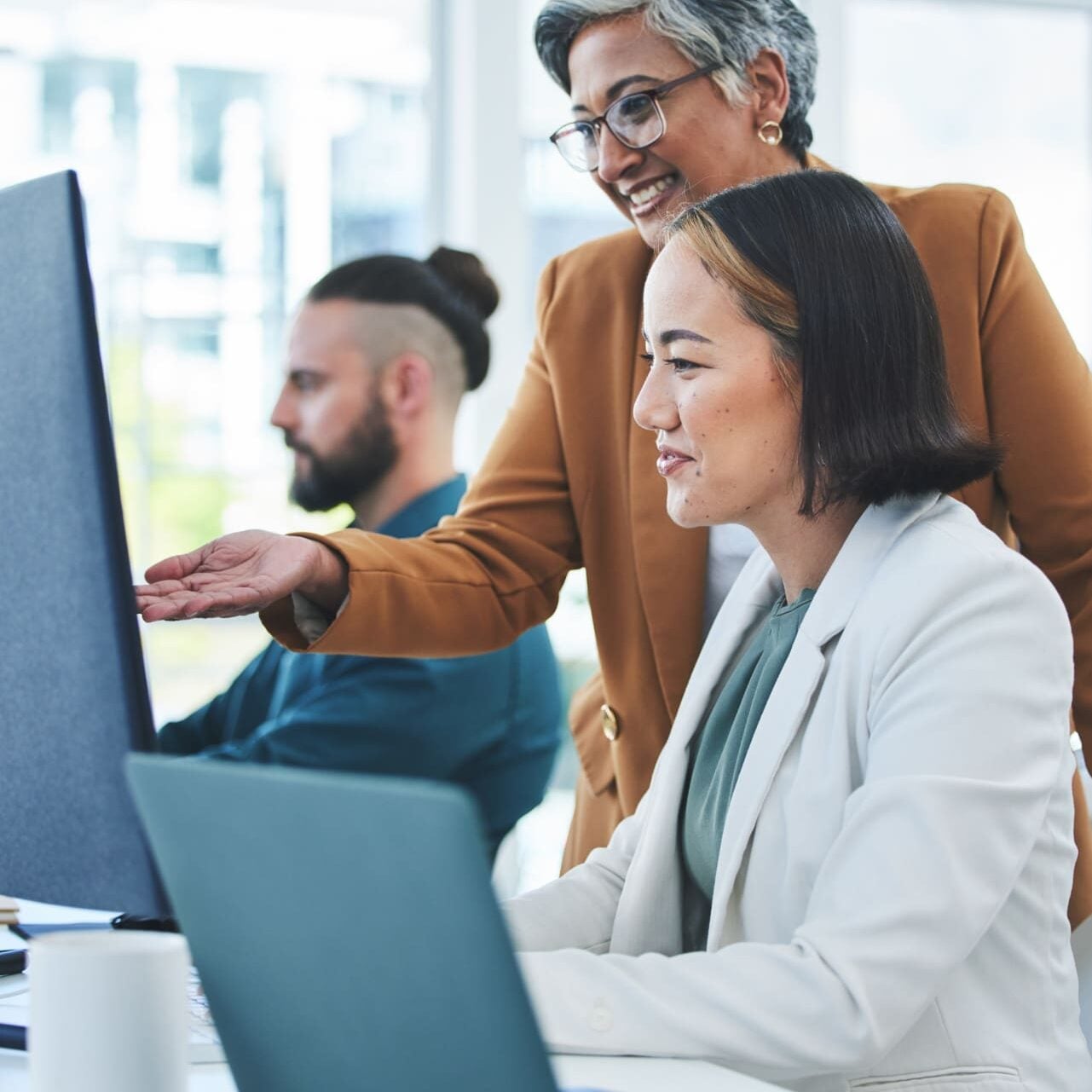 Two women in business attire smile and look at a computer screen together in an office, while a man works at a desk in the background. The atmosphere is collaborative and professional.